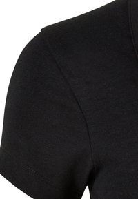 Gros plan sur l'épaule et la couture de la manche d'un T-shirt en coton noir à manches courtes avec des coutures visibles sur un fond blanc uni.