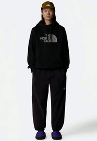 Νεαρός άνδρας που φοράει μαύρη ζακέτα και παντελόνι The North Face, ελιάς πράσινο καπέλο και μοβ παντόφλες, στέκεται μπροστά σε λευκό φόντο.