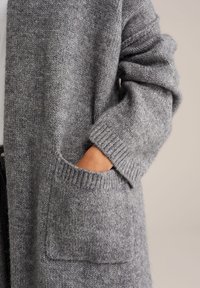 Pelēks adīts kardigans ar mīkstu, teksturētu audumu, ar kabatu priekšpusē un ribotām piedurknēm, demonstrējot brīvu piegriezumu.