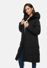 Long black puffer coat with a hood, featuring quilted texture, front zip closure, and side pockets. Model stands with hands on the collar.