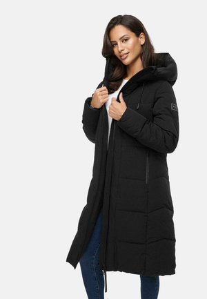 Lange zwarte puffercoat met capuchon, voorzien van een gequiltde textuur, een rits aan de voorkant en zijzakken. Model staat met de handen op de kraag.