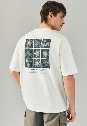T-shirt en coton blanc avec un graphisme carré au dos présentant neuf illustrations florales et d'étoiles en vert foncé, accompagnées d'un texte blanc en dessous.