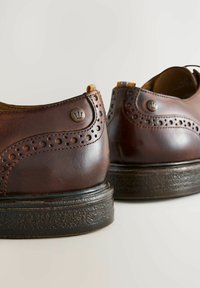 Chaussures en cuir marron avec des perforations décoratives, accents de logo rond et semelles texturées. Présente un design classique et une finition lisse.