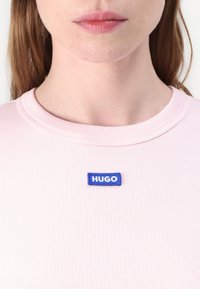 Nærbillede af en person iført en lyserød ribbet skjorte med et blåt rektangulært mærke, hvor der står "HUGO" i centrum af halsudskæringen.