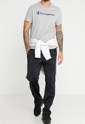 Muž nosící šedé tričko Champion, bílou mikinu uvázanou v pase, černé kalhoty s bočními pruhy a černé tenisky, stojící s rukama v kapsách.