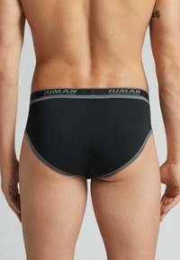 Schwarze Herrenunterwäsche aus glattem Stoff, ausgestattet mit einem grauen Bund mit Logo und kontrastierenden Verzierungen. Der Schnitt bietet minimale Bedeckung am Rücken.