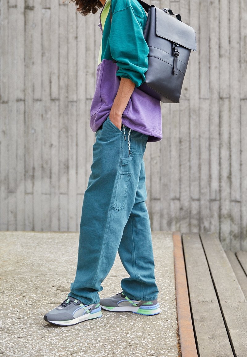 Colorful oversized hoodie in teal and purple, relaxed-fit teal pants, and gray-green sneakers with blue accents; black backpack with flap.