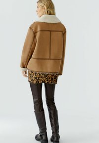 Veste en daim marron avec un col en fausse fourrure, présentant un ourlet à motif léopard. Assortie avec un legging en cuir marron et des bottes à hauteur de genou.