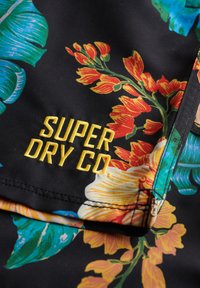 Tejido con estampado floral de colores vibrantes, con grandes hojas y flores tropicales. Texto bordado en amarillo "SUPER DRY CO". Fondo negro.