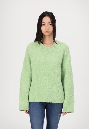 Hellgrüner, strukturierter Strickpullover mit V-Ausschnitt und langen Ärmeln, in lässiger Passform und mit geripptem Saum, kombiniert mit blauen Jeans.
