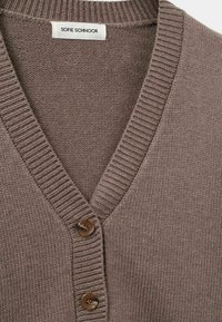 Sveter s V-výstrihom vyrobený z mäkkého textúrovaného hnedého pleteného materiálu, ktorý má rebrované detaily a dva hnedé gombíky vpredu.