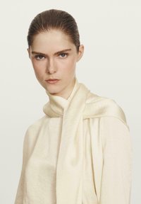 Crème-kleurig kledingstuk met een zachte, textuurrijke stof en een gedrapeerd sjaaldetail rond de hals, wat een soepel silhouet creëert.