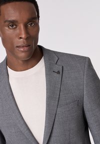 Grau strukturierter Blazer mit dezentem Hahnentrittmuster, ausgestattet mit Reverskragen und einer Brusttasche. Kombiniert mit einem hellbeigen Rundhalsausschnitt-Pullover.