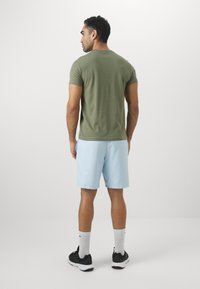 Olivno zeleni kratkolisni T-shirt v kombinaciji z svetloblušimi kratkimi hlačami. Kratke hlače so ohlapnega kroja z gladko teksturo. Črne športne čevlje in bele nogavice.