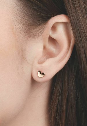 Primer plano de una oreja izquierda con cabello castaño liso recogido detrás, que lleva un pequeño pendiente tipo tachuela pulido en forma de corazón dorado.