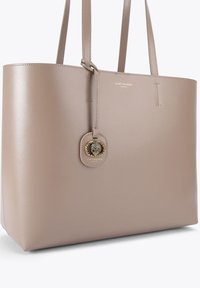 Beżowa torba typu tote z syntetycznej skóry o gładkiej fakturze, z dwoma paskami na ramię i złotym medalionem z logo. Przestronny prostokątny kształt.