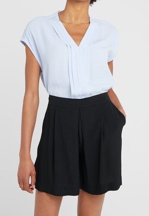 Frau trägt eine hellblaue V-Ausschnitt-Bluse mit Falten, in hoch taillierte schwarze, plissierte Shorts gesteckt, Hand in der Tasche vor einfarbigem Hintergrund.