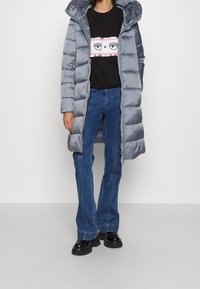 Persona che indossa un lungo piumino grigio, felpa nera con stampa di occhi, jeans svasati blu e scarpe nere spesse su uno sfondo neutro.