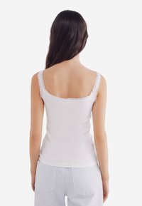 Valkoinen camisole, jossa on pitsiset hihnat, istuva muotoilu, ribbineule pinta ja pyöreä pääntie, yhdistettynä vaaleisiin raidallisiin housuihin.