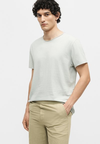 Νεαρός άνδρας με σγουρά σκουρόχρωμα μαλλιά, φοράει απλό ανοιχτό γκρι μπλουζάκι και μπεζ παντελόνι με το ένα χέρι στην τσέπη.