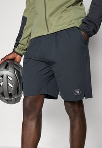 Donkergrijze shorts met een gladde textuur, voorzien van zijzakken en een logo-opdruk, gedragen met een groene jas en een grijze helm in de hand.