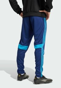 Persona vistiendo pantalones deportivos azules con rayas azul claro y zapatos de fútbol negros, de pie con las manos en las caderas, de espaldas.