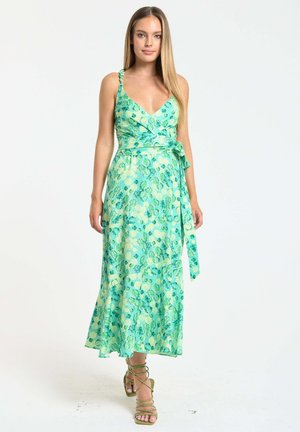 Woman wearing a sleeveless green floral wrap dress with a side tie, walking forward in strappy high-heeled sandals on a white background.