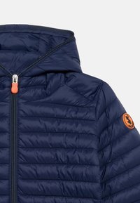 Ναυτικό μπλε καπιτονέ μπουφάν με κουκούλα, πορτοκαλί φερμουάρ και κυκλικό πορτοκαλί έμβλημα στο πάνω μέρος του μανικιού.