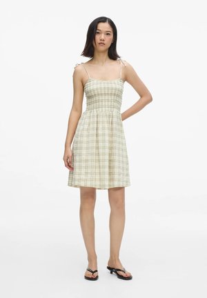 Femme portant une robe beige à carreaux sans manches avec des bretelles à nouer, debout pieds nus sur un fond blanc, face à l'avant avec une main sur la hanche.