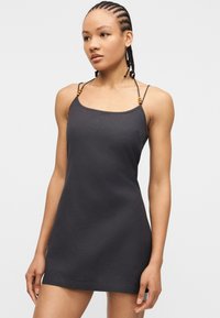 Νεαρή γυναίκα με πλεξούδες, που φορεί ένα μίνι μαύρο φόρεμα χωρίς μανίκια με λεπτούς ιμάντες και διακοσμητικά από ξύλινες χάντρες, στέκεται μπροστά σε λευκό φόντο.