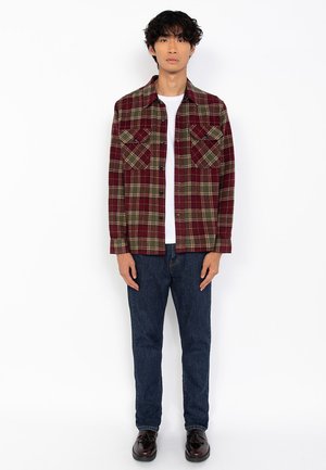 Jeune homme debout, portant une veste en plaid rouge et vert sur une chemise blanche, un jean bleu foncé et des mocassins à glands marron foncé.