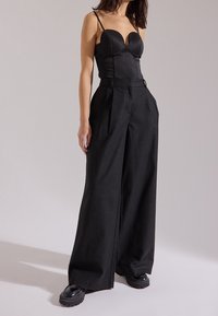 Donna che indossa un top bustier attillato nero con spalline sottili, pantaloni neri a vita alta e larghi, e scarpe nere chunky, in piedi di fronte a uno sfondo chiaro e semplice.