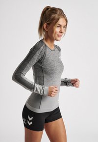 Ung kvinna som joggar i grå långärmad träningsöverdel och svarta shorts mot en enfärgad vit bakgrund.