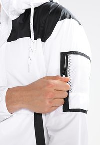 Herre vindjakke i hvid og sort, med hætte, kontrasterende skulderdesign og en lynlåslomme i mesh på armen.