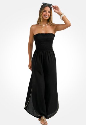 Kobieta w czarnym, bezramkowym kombinezonie z szerokimi nogawkami i rozcięciami po bokach wykończonymi złotą lamówką, w towarzystwie naszyjnika z muszli, bransoletek i sandałów.