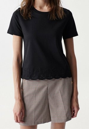 Femme portant un haut noir à manches courtes avec des découpes florales à l'ourlet et un short à carreaux taille haute avec des boutons sur les côtés.