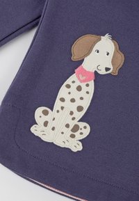 Purple fabric featuring an embroidered beige dog with brown spots, wearing a pink collar, on the front of a garment. лапка