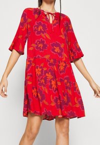 Puna mekko, jossa on violetti ja oranssi kukkakuosi, kerroksellinen muotoilu, lyhyet röyhelösleeve-t ja avonainen kaula-aukko, jossa on nauhakiristys.