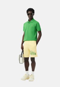 Camisa polo verde, pantalones cortos amarillos con gráficos verdes, zapatillas blancas y una bolsa blanca con detalles negros. La tela es casual y ligera.
