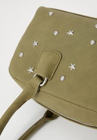 Sac en suédine verte olive avec des embellissements en étoile argentée et des clous, fermeture à glissière et une bandoulière plate pour le transport. Surface texturée.