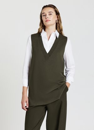 Gilet sans manches vert olive avec un décolleté en V profond, superposé sur une chemise blanche à boutons. Texture lisse, coupe décontractée, détails minimaux.