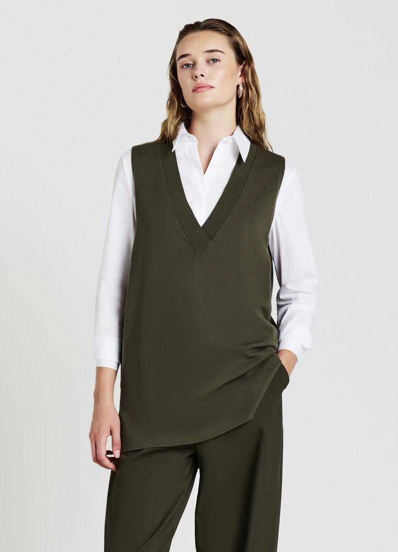 Maglione senza maniche verde oliva con scollo a V profondo, indossato sopra una camicia bianca a maniche lunghe. Texture liscia, vestibilità comoda, dettagli minimi.