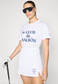 Weißes T-Shirt mit blauer bestickter Aufschrift "le CLUB du MALBÔN", kombiniert mit einem weißen Golfrock. Das Model trägt schwarze reflexive Sonnenbrille und einen Golfhandschuh.