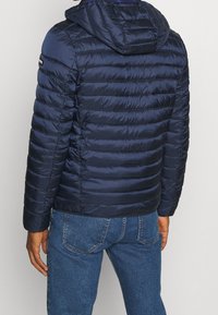 Mørkeblå dunjakke med hætte, med quiltede sektioner for isolering, glat tekstur og en lille logo på skulderen.