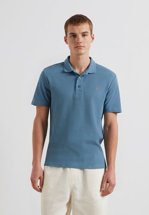 Junger Mann, der ein blaues kurzärmeliges Poloshirt und hellbeige Hosen trägt, steht vor einem schlichten weißen Hintergrund.