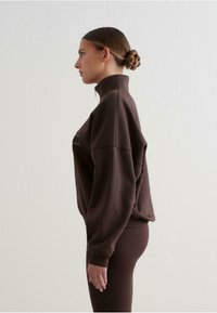 Sudadera de forro polar marrón con cuello alto, corte relajado y puños acanalados. Presenta texto de logo en el frontal, combinada con leggings marrones a juego.