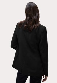 Svart skräddarsydd kavaj med en strukturerad design, slät tyg och något rundad bakre fåll. Ärmarna är åtsittande med en avslappnad silhuett.