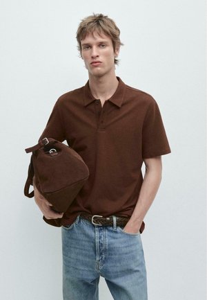 Jeune homme portant un polo marron et un jean bleu, tenant un sac de voyage en cuir marron d'une main, l'autre main dans sa poche.