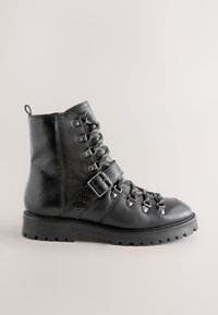 Musta nahkainen nilkkuri, jossa on teksturoitu sivu, metalliset reiät ja nauhoitusjärjestelmä. Paksu pohja ja solkikoriste hihnassa.