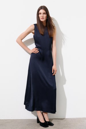 Kvinne med langt brunt hår står mot en hvit vegg iført en ermeløs marineblå midi-kjole og svarte sko med blokkhæl.